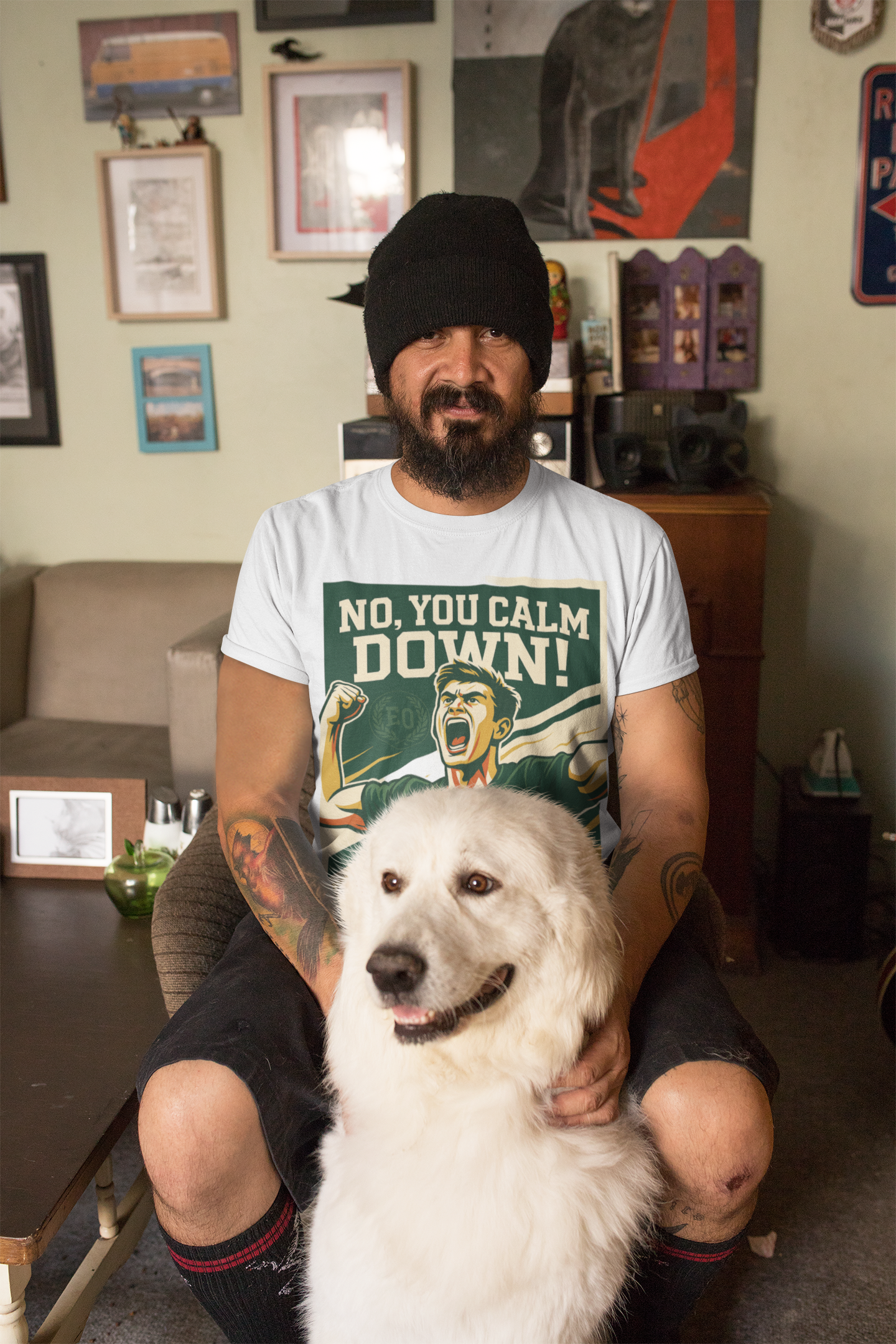 Man wearing a t-shirt with a graphic design and text, sitting with a white dog in a room with various items on the walls.