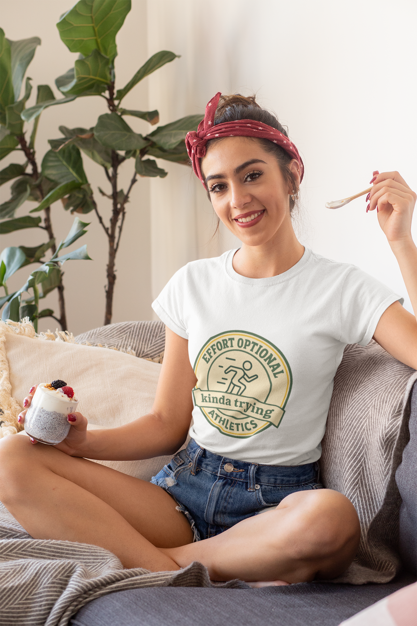 Woman sitting on a couch holding a cup and a spoon, wearing a white t-shirt with a graphic design.