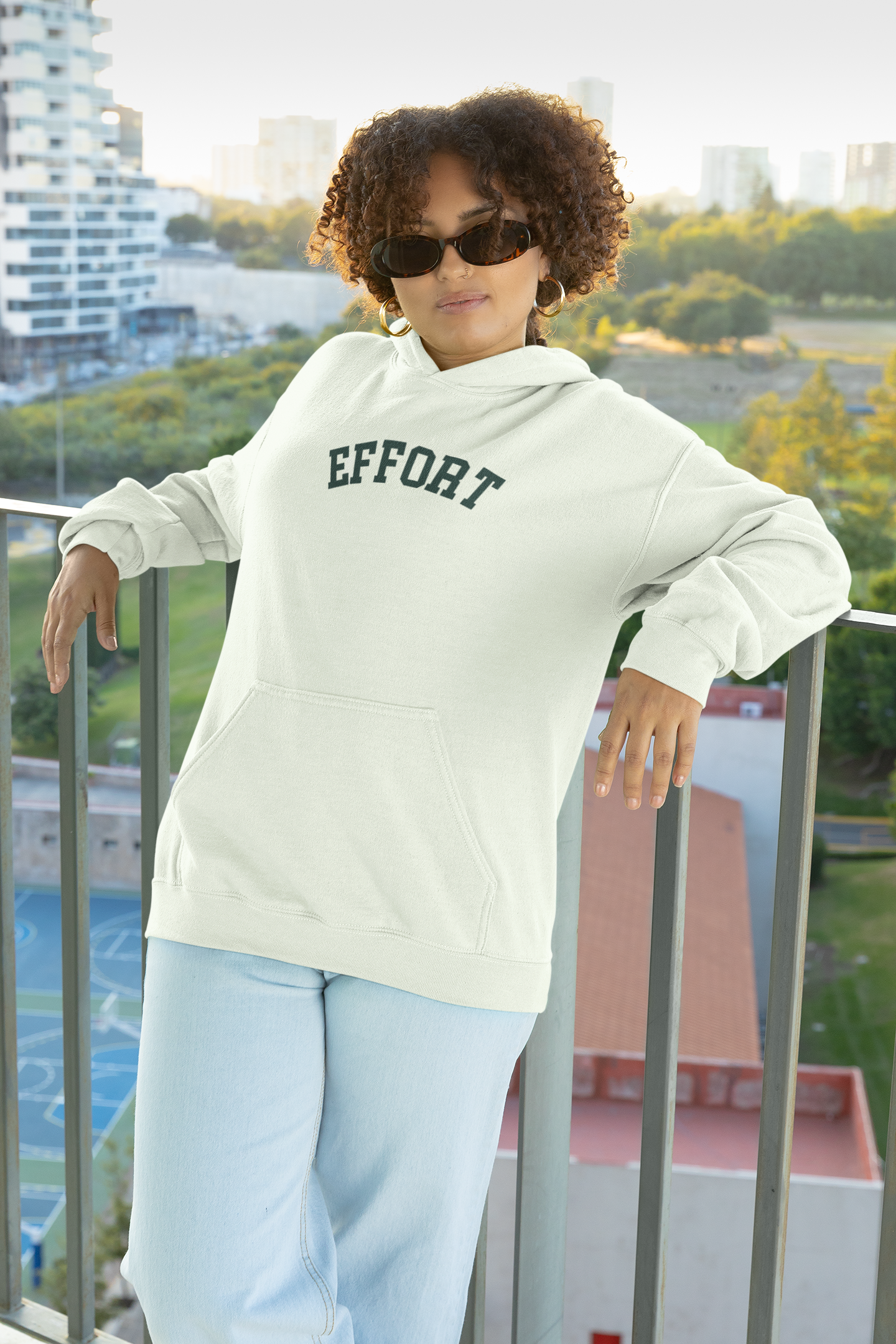Person wearing a light-colored hoodie with 'EFFORT' text, standing on a balcony with a cityscape background.