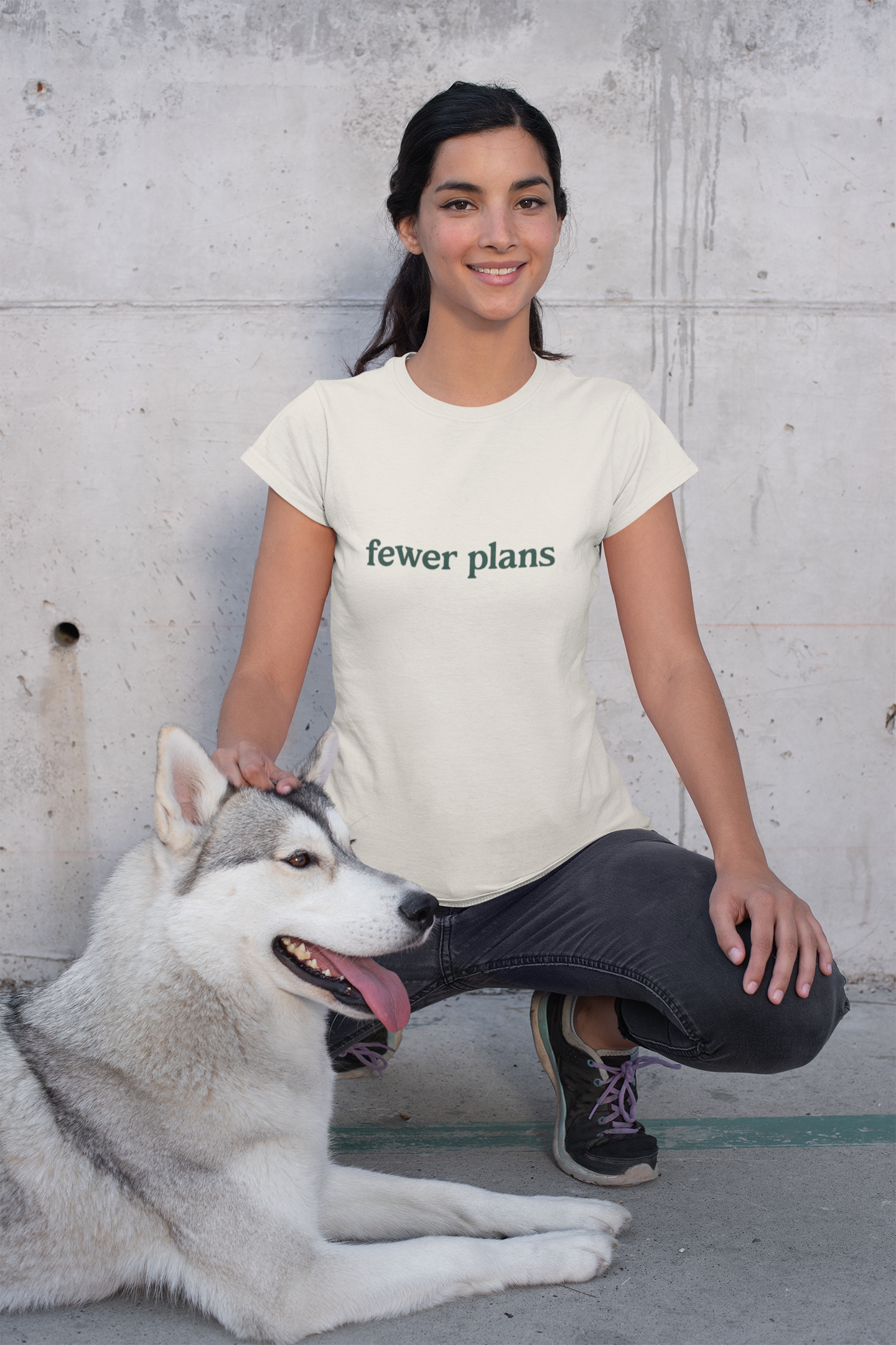 Woman wearing a 'fewer plans' t-shirt sitting with a dog against a concrete wall.