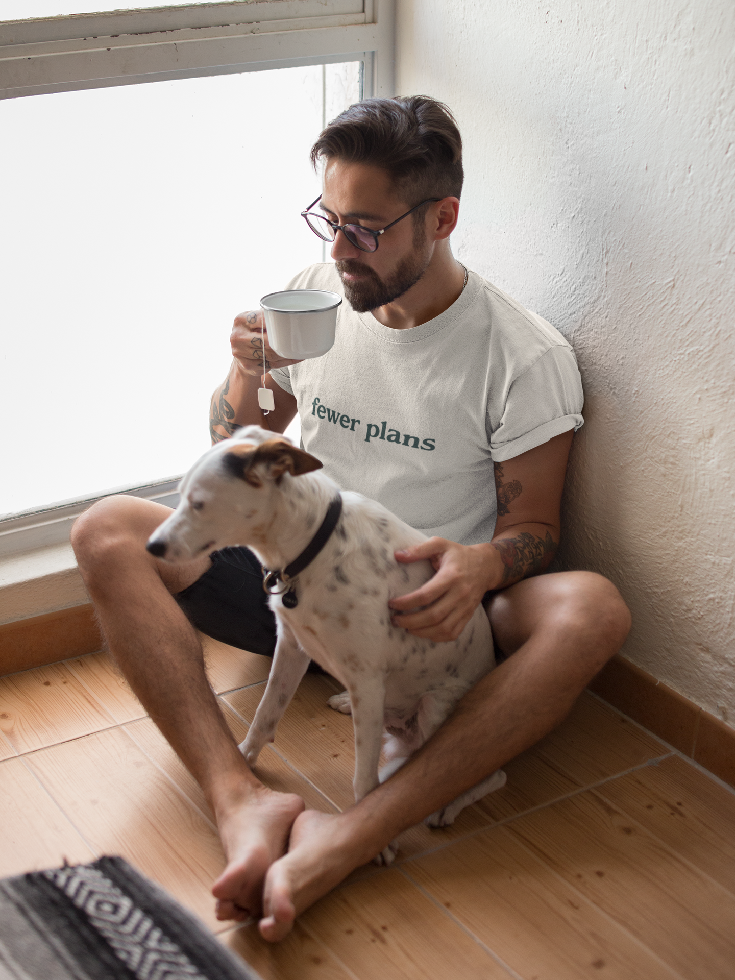 Man sitting on the floor with a dog, holding a mug by a window.