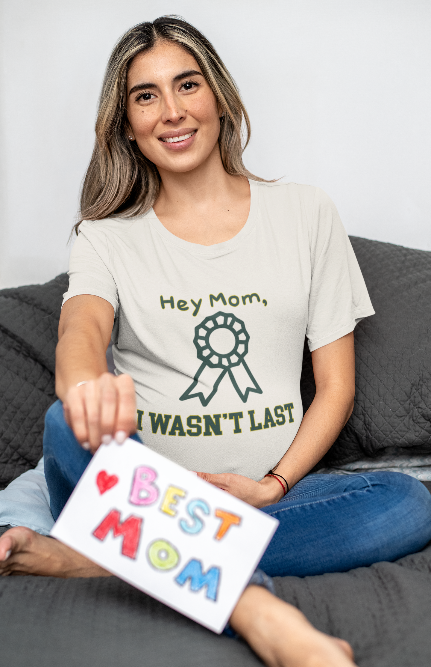 Woman wearing a t-shirt with 'Hey Mom, I wasn't last' text, sitting on a couch holding a sign saying 'Best Mom'.