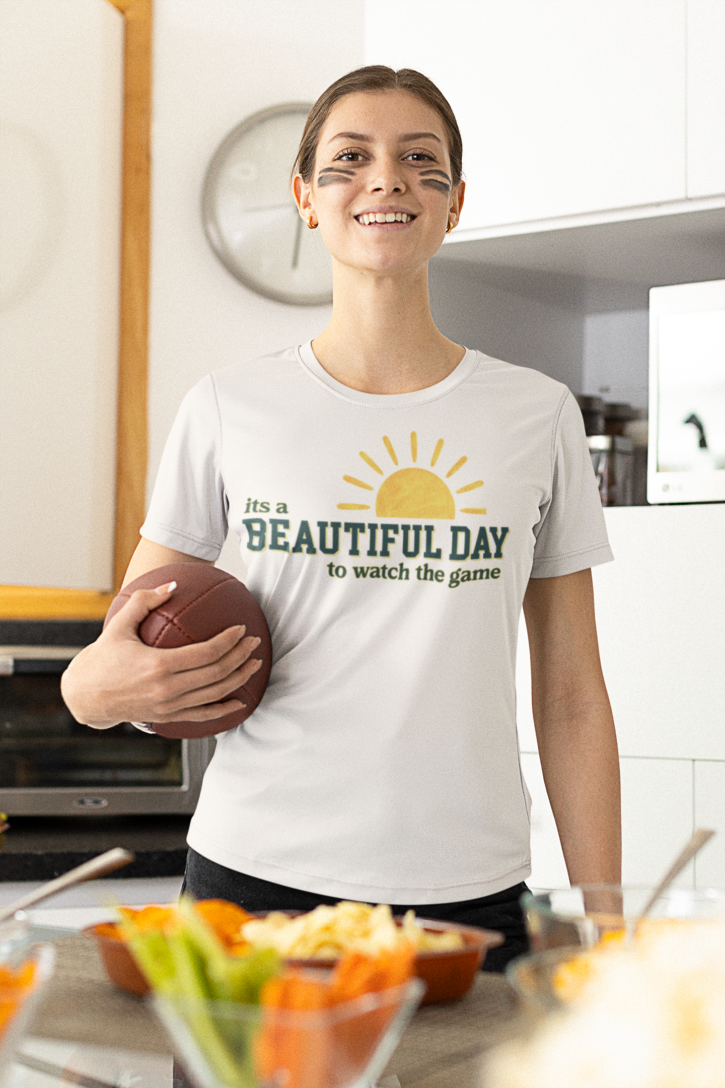 Woman wearing a 'It's a Beautiful Day to watch the game' t-shirt in a kitchen.