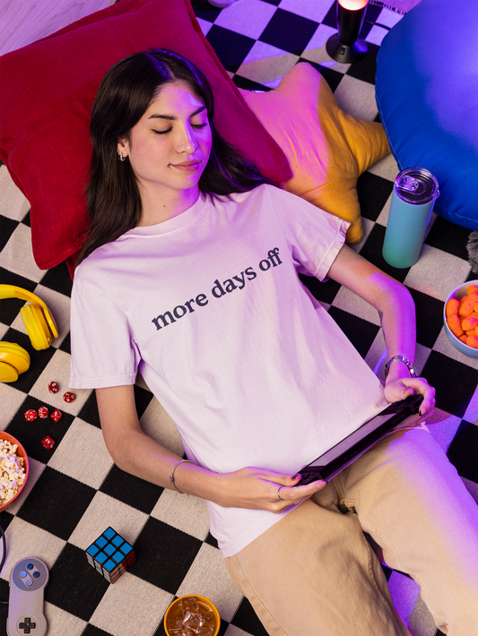 Woman lying on a checkered floor wearing a white t-shirt with text, surrounded by colorful pillows and snacks.