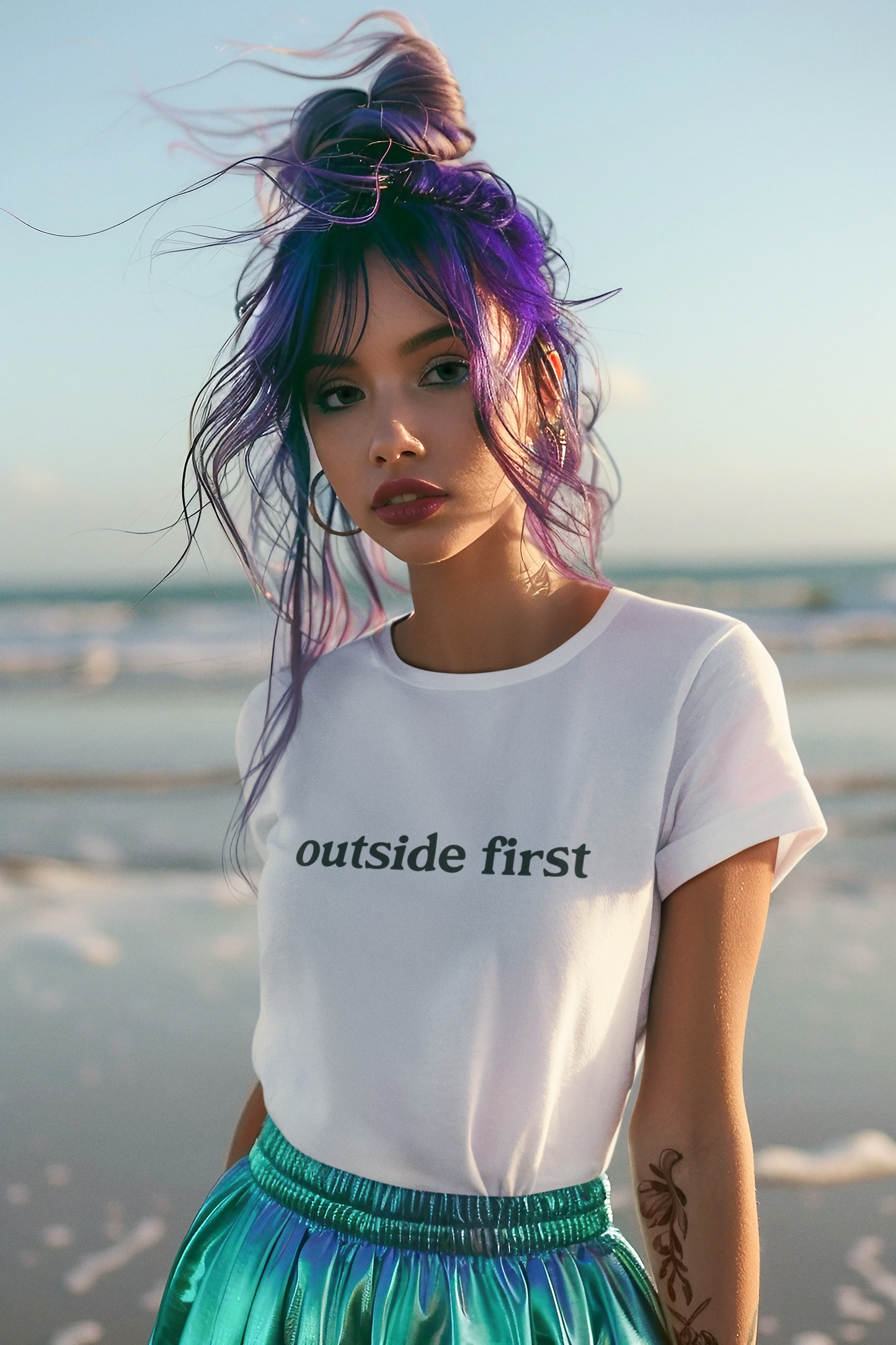 Person with purple hair wearing a white t-shirt with 'outside first' text on a beach.