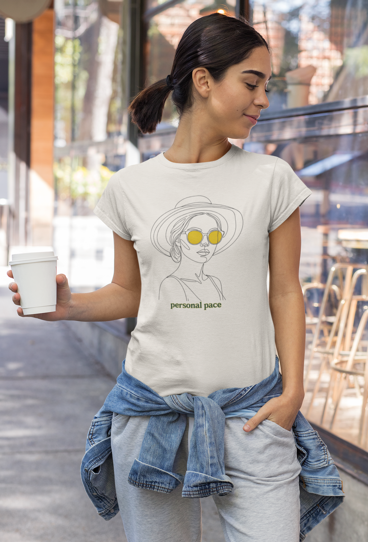 Woman wearing a t-shirt with a graphic design and text, holding a cup outdoors.