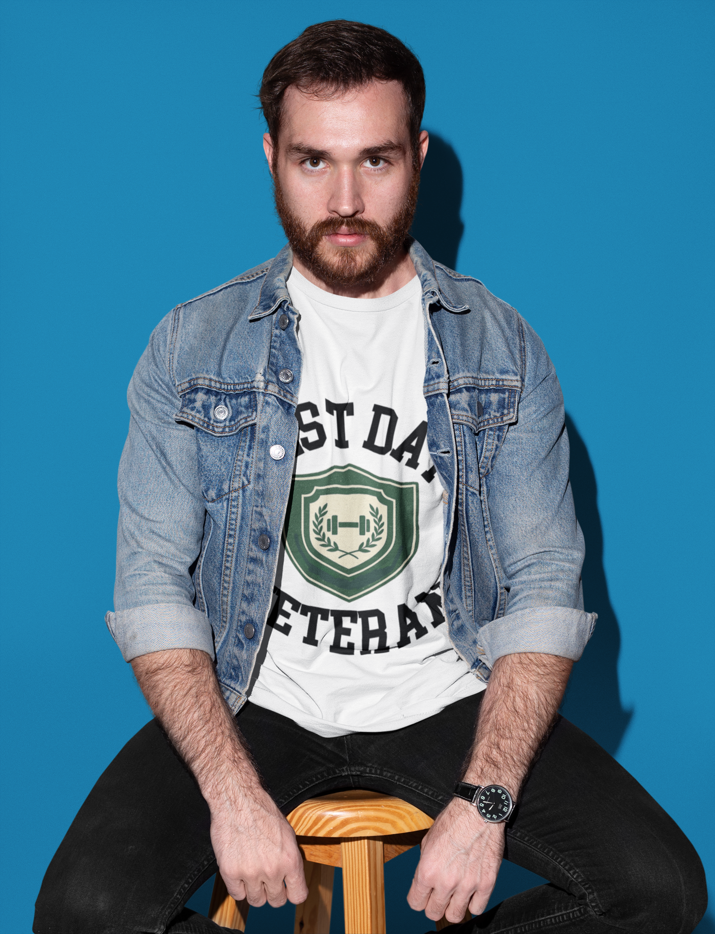 Man wearing a denim jacket over a white t-shirt with text and graphics, sitting on a stool against a blue background.