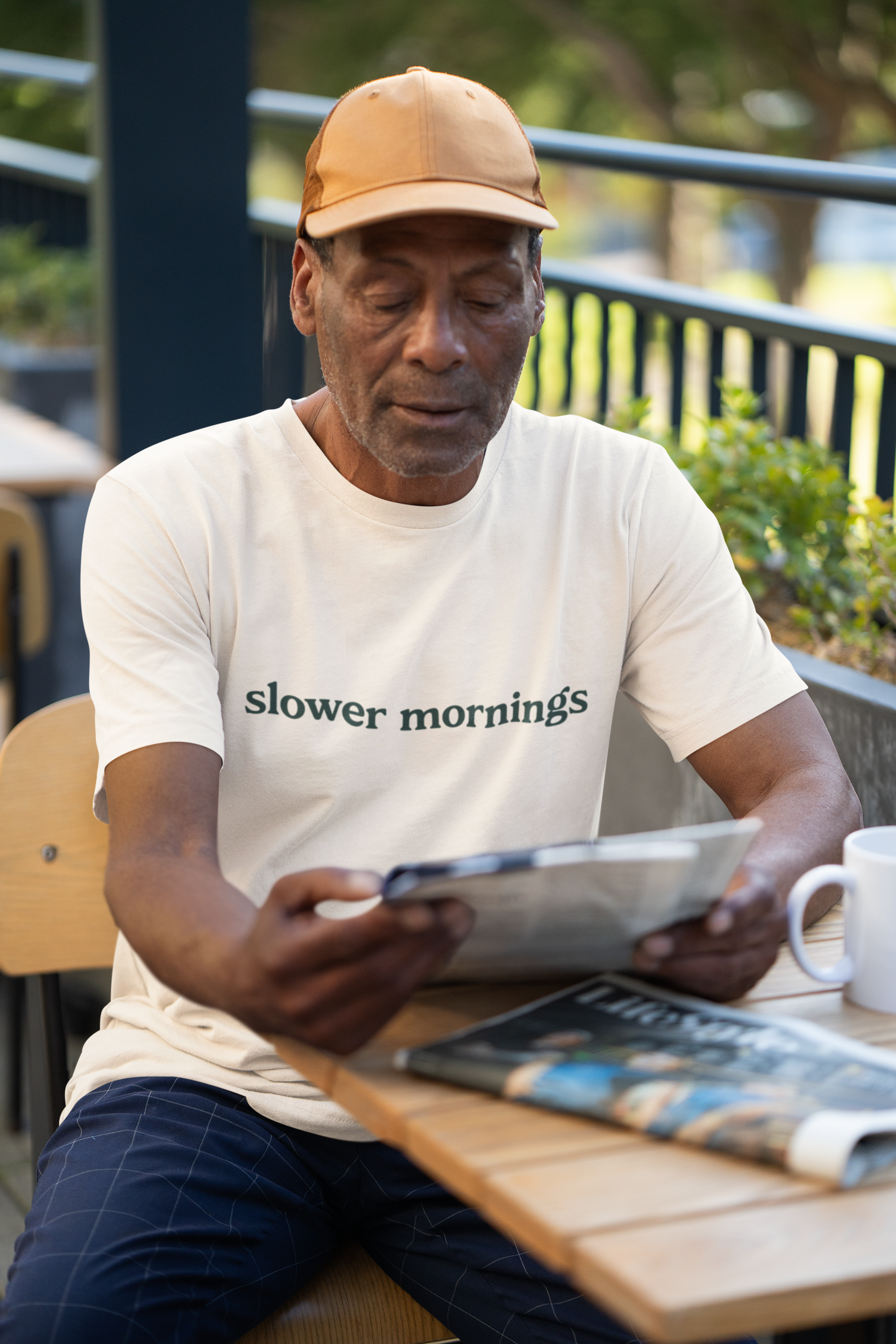 Man reading a newspaper outdoors with a cup of coffee and magazine on a table.