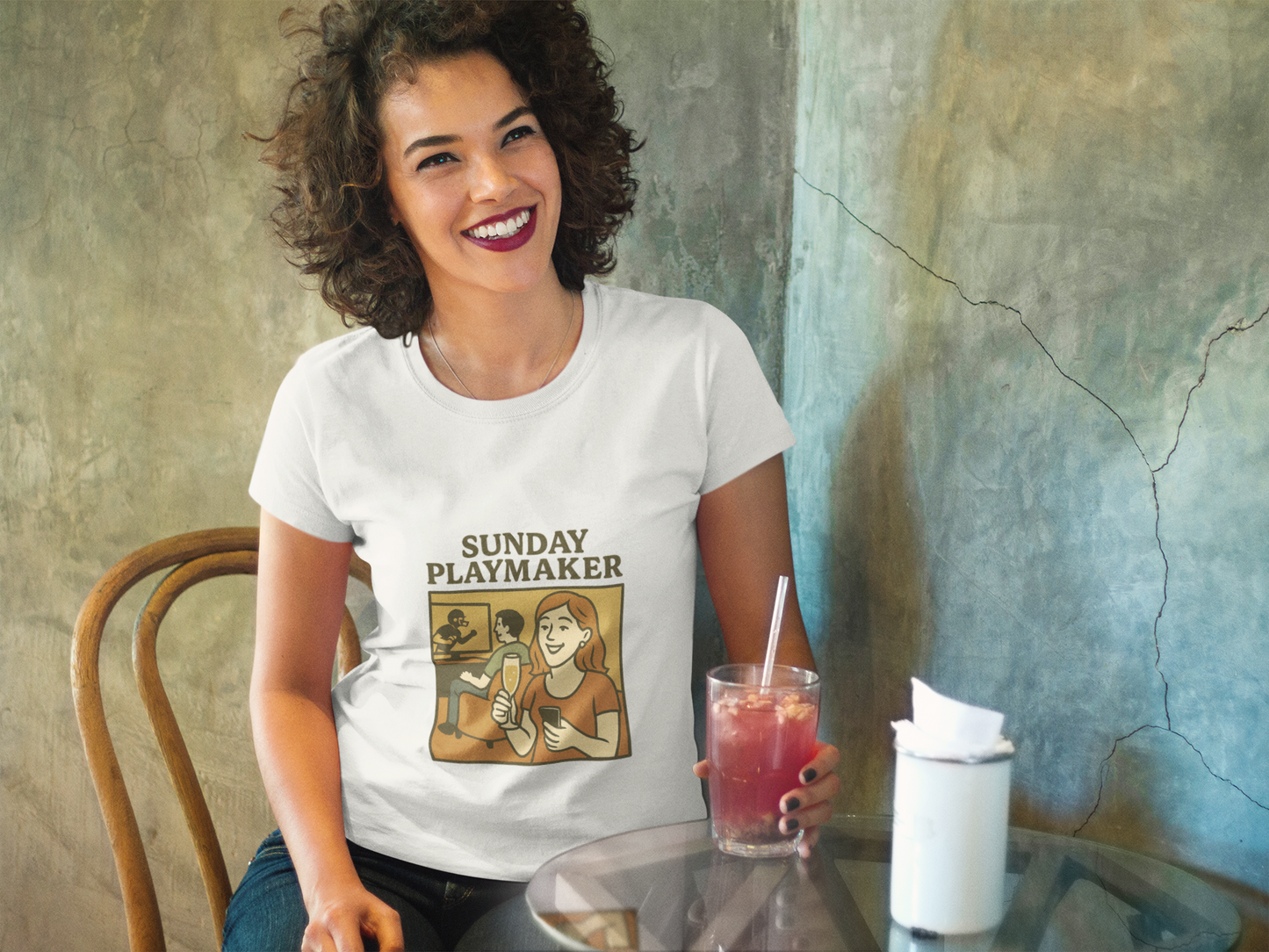 Woman wearing a 'Sunday Playmaker' t-shirt sitting at a table with a drink.