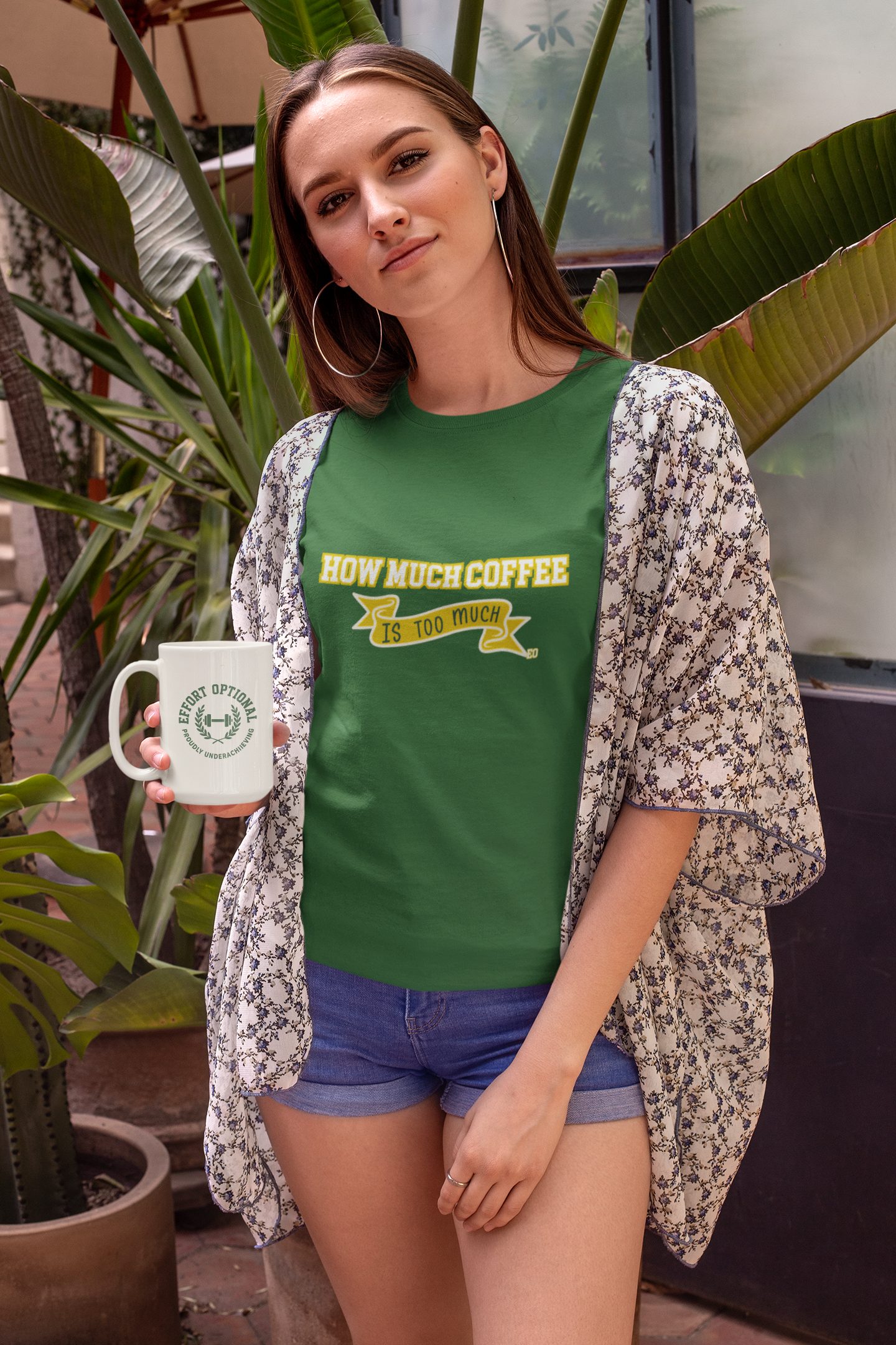 Woman wearing a green t-shirt with text, holding a mug, standing outdoors with plants in the background