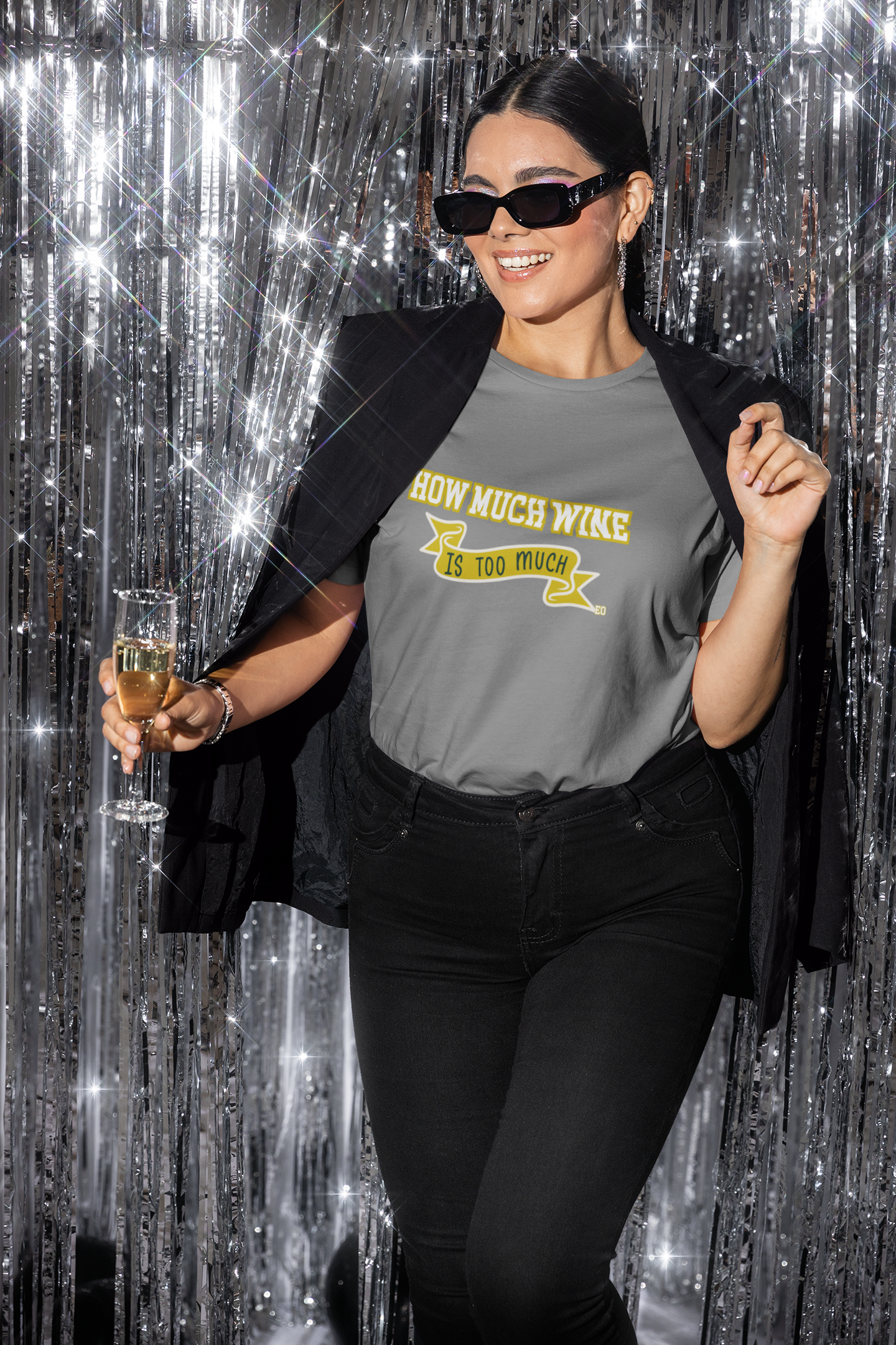 Woman in a gray t-shirt with text, black pants, and sunglasses holding a glass against a silver curtain background.