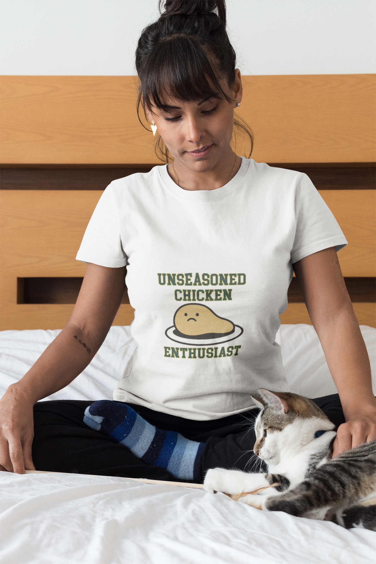 Woman wearing a t-shirt with 'Unseasoned Chicken Enthusiast' graphic sitting on a bed with a cat.