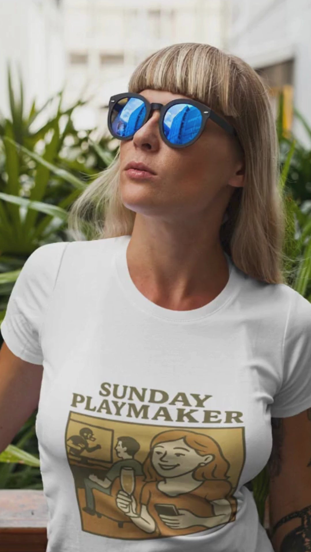 Woman wearing a white t-shirt with 'Sunday Playmaker' graphic design, standing outdoors with plants in the background. Video